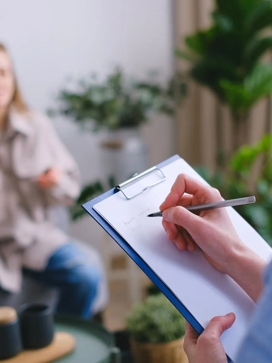 Clinician taking notes while a patient speaks in a warm, plant-filled therapy room
